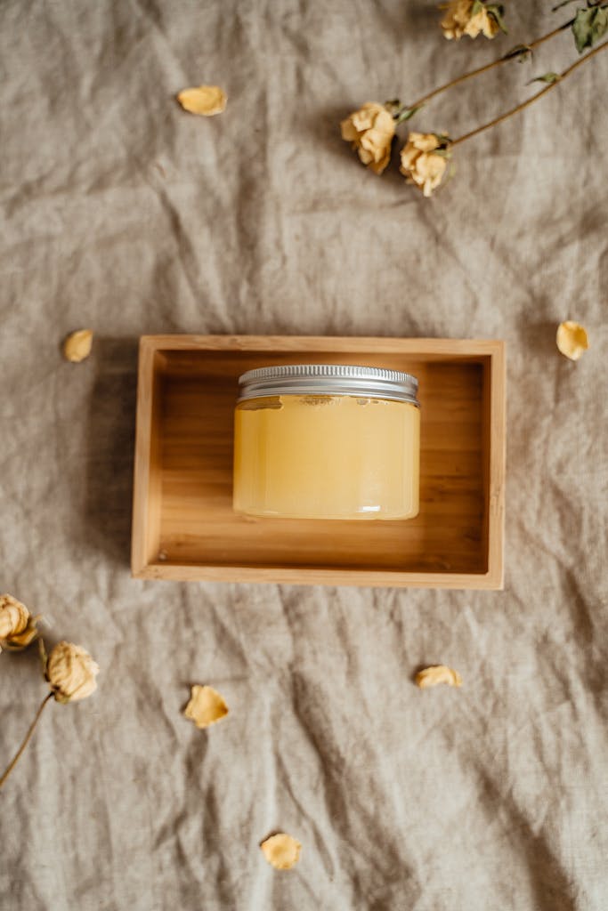 A glass jar of yellow skincare product displayed in a wooden box with dried flowers.