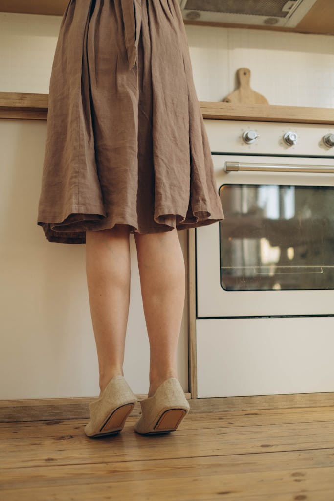 A woman in a beige dress tiptoes on heels in a warm, cozy kitchen setting.