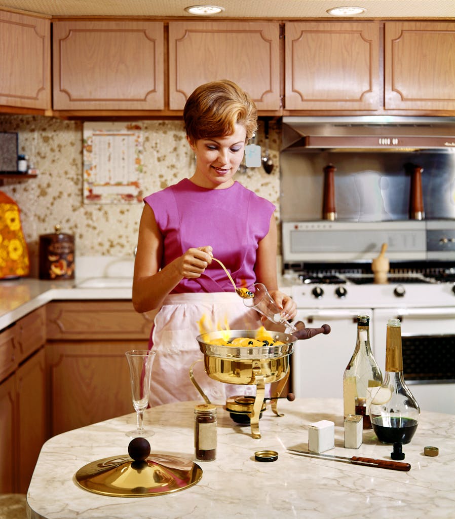 Vintage kitchen scene with a woman cooking a flambé dessert, showcasing retro style and gourmet preparation.