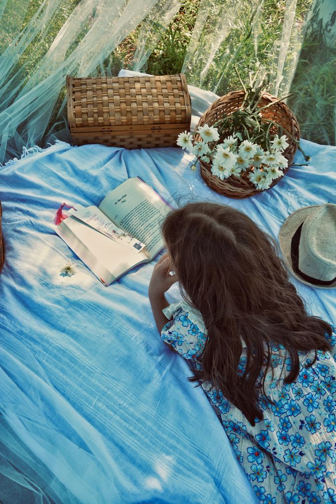 Young girl reading a book on a picnic blanket outdoors surrounded by flowers and basket.