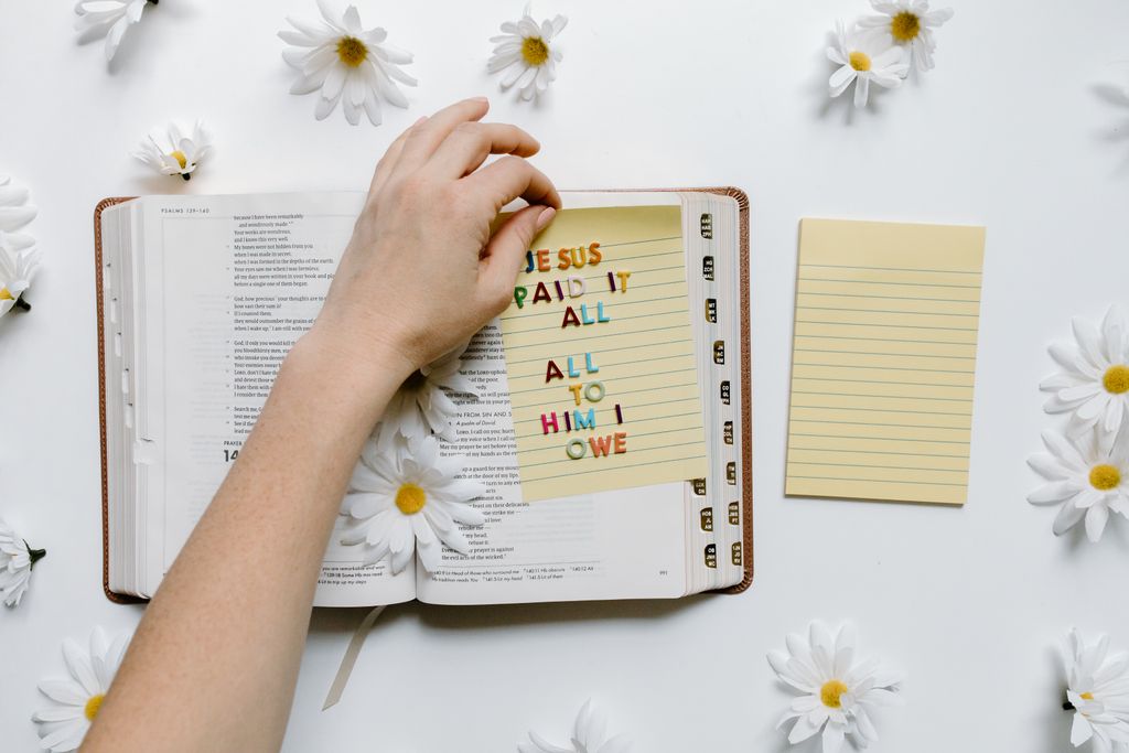 A hand holding a bright message card on an open Bible surrounded by daisies.