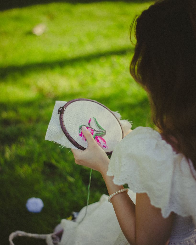 A woman in a white dress sewing a floral pattern in a sunny, grassy park setting.