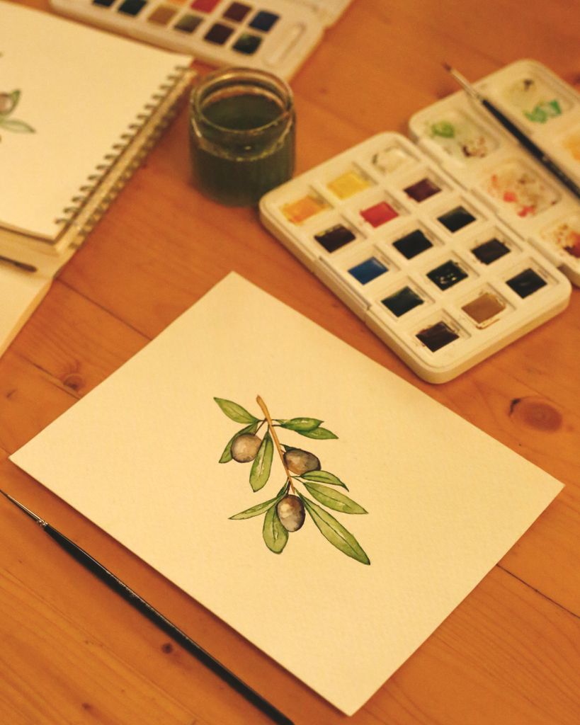 Elegant watercolor of olive branch next to art supplies on a wooden table.