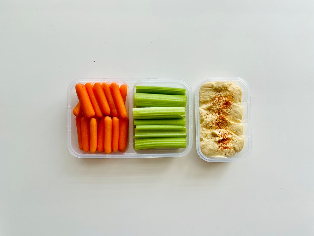 Fresh baby carrots, celery sticks, and hummus in a container for a healthy snack.