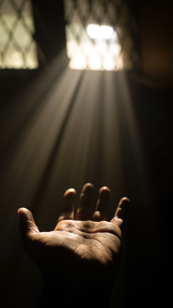 A single hand reaches out towards a soft beam of light streaming through a window.